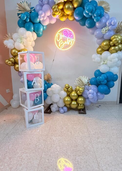 Circular balloon arch in white, lavender, teal, and gold with a glowing “Oh Baby” neon sign and a birthday balloon box setup of transparent stacked boxes spelling “BABY” filled with balloons.