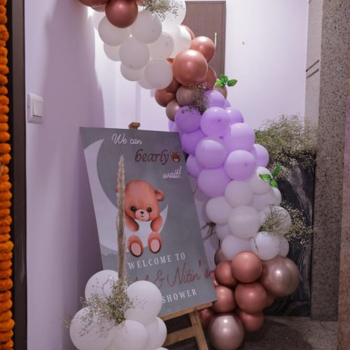 Baby shower balloon arch with white, rose gold, and light purple balloons beside a welcome sign with a teddy bear illustration in an indoor hallway.