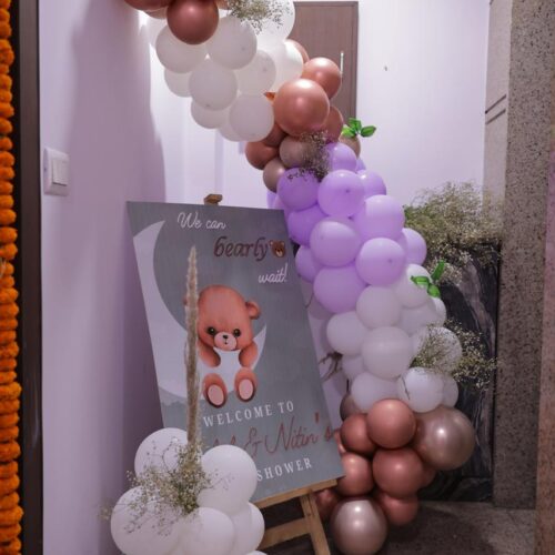 Baby shower balloon arch with white, rose gold, and light purple balloons beside a welcome sign with a teddy bear illustration in an indoor hallway.